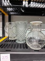 Three pitchers displayed on shelf, two vintage pressed cut-glass and one clear glass pitcher with white lid ice container.