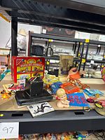 Photo showing a collection of vintage toys, cards, buttons, and small memorabilia on a display shelf including a porcelain Kewpie doll, Earldums Animals card game tin, various colorful buttons, a small black box, and several small plastic toys.