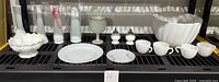 Display of white milk glass serveware including vases, plates, salt and pepper shakers, punch bowl with cups.