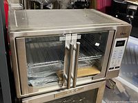 Front and side views of Oster digital countertop convection oven showing stainless steel finish, double glass doors with handles, digital control panel with display and buttons, and interior items including a baking tray wrapped in plastic and metal rack.