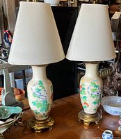 Pair of Asian-inspired porcelain table lamps with painted floral design, cream fabric lampshades, standing on brass bases with minor wear.