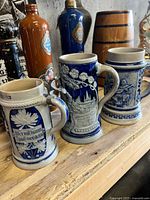 Set of three German stoneware beer steins with blue and grey relief designs and German text inscriptions. All steins shown without lids, placed on wooden surface.