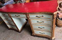 Full angled view of vintage writing desk showing multiple drawers painted in muted greenish-blue, red curvy top surface, and gold-toned ornate handles.