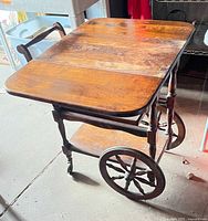 Side angle showing the drop-leaf sides partially expanded, large wooden spoked wheels, turned legs, and lower shelf with handle cutout.