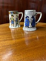 Two Royal Doulton ceramic tankard mugs placed on a wooden surface, showing front views with detailed painted figures, one in blue tones and one in colorful cavalier attire.