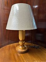 Full view of the wood turned table lamp with new light tan fabric shade placed on round wooden base, cord coiled beside it.