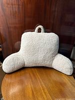 Front view of light beige high pile fluffy back rest cushion with carrying handle at top center, placed on wooden surface against dark wood background.