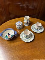 Full set of Henriot Quimper coffee service including milk pitcher, covered sugar bowl, two coffee cans with saucers, and Isabelle porringer displayed on a wooden table.