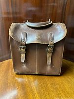 Front view of antique brown leather lawn bowling bag with double buckle straps and flap, showing gold initials 'A.S.' on the flap.