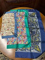 All three Liberty silk scarves laid overlapping on a wooden table, visible patterns include blue paisley, green border with flowers, and steel blue border with cherries and leaves.