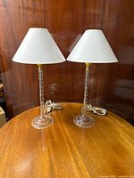 Pair of clear crystal lamps with white fabric shades on a wooden table.