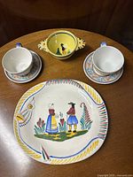 Full display of serving plater with painted figures, two tea/coffee cups with saucers, and yellow porringer.