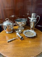 Full set of six silver plated barware pieces on wooden table, including punch bowl, ice bucket, cocktail shaker, jigger, two wine coasters.