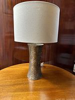 Full view of the modern table lamp showing its textured silver to bronze base and white fabric shade on a wooden surface with a wooden background.