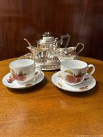 Three-piece silver plated EPNS tea set displayed on the tray with two Royal Worcester Evesham cups and saucers in front, showing clear fruit and floral decoration on the china and gadroon silver detailing on the plated items.