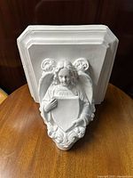 Front view of white painted wall bracket showing angel holding shield with detailed wings and facial features.