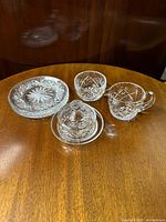 Photo showing creamer, sugar bowl, butter dish with lid, and three dessert/salad plates arranged on a wooden surface illustrating the full set and cut crystal detail.