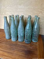 14 old Coca-Cola glass bottles grouped together showing different greenish and clear glass bottles.