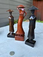Three carved teak Mid-Century Modern style geisha statues placed on outdoor surface with coin for scale.