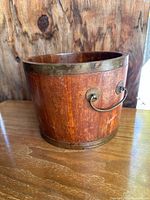 View of the wooden bucket showing brass handle and brass band on top and bottom rim, highlighting the wood grain and antique finish