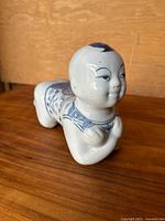 Front-side perspective of antique blue and white porcelain opium pillow shaped like a child lying on stomach with arms crossed.
