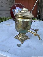 Mini brass Russian samovar with lid, spigot and pedestal base seen on a white outdoor surface, showing the overall shape and condition.
