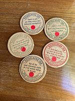 Photo showing all five antique Heidelberg Inn coasters arranged on a wooden surface, each with distinct witty sayings visible and red circular stickers with numbers on them.