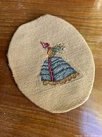 Close-up of the handmade crossstitch embroidery showing the full figure of the woman in a Victorian dress with a bonnet and holding flowers on beige fabric.