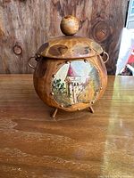 Front view showing the wooden carved jar with a painted village or castle image on its side, spherical lid knob, and three short legs supporting it.