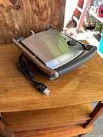 Stainless steel Breville panini press showing closed top and cord neatly wrapped beside the unit on a wooden surface.