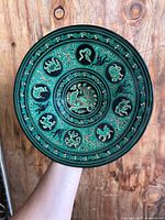 Full view of carved lacquer plate held vertically against wood background showing detailed central phoenix design and surrounding mythical figures.