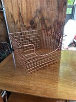 Copper-colored wire basket with open grid wire mesh, placed on wooden surface with wood wall backdrop, showing front and side views.