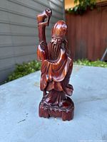 Front view of the hand carved teak Chinese elder statue on a surface with background of wooden wall and greenery.