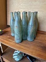Group of 10 greenish antique Coca-Cola bottles arranged on wooden shelf, showing general condition and grouping.