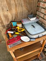 Image showing a wooden shelf with a variety of kitchen utensils spread out including a plastic storage container with lid, a white spoon rest, and multiple peelers and slicers.