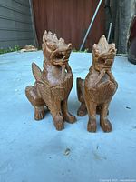 Pair of carved wooden foo dogs pictured on a blue surface outdoors against a dark wood wall, showing both statues standing side by side.