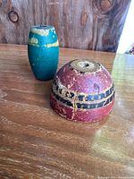 Pair of antique wooden fishing floats on wooden surface against rustic wooden backdrop. Green cylindrical float with yellow band and red dome-shaped float with black and gold band visible.