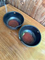 Pair of black glazed ceramic bowls with reddish brown interior, each with a chopstick rest notch and hole on the rim, placed on wood surface.