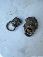 Two metal lion head door knockers side by side on a flat surface. Each knocker is a lion's head holding a round metal ring in its mouth. Visible patina and wear on the metal.