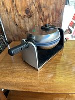 Side angle view of stainless steel waffle maker with black handle and base on wooden surface.