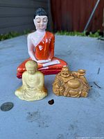 Three Buddha figurines arranged on a flat surface outside with house siding in background; a medium-sized painted Buddha in orange robe, small beige Buddha and laughing Buddha figurine.