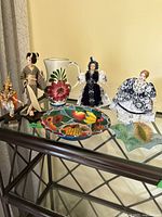 Asian and European dolls grouped with Spanish ceramic water jug and colorful Cuban paper art bowl on glass table.