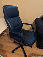 Blue leather office chair angled left side showing seat, backrest, and armrest on wood floor near white table and dark couch.