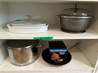 Shelf with CorningWare white ceramic rectangular baking dish with clear glass lid on top shelf; stainless steel pot with lid, black frying pan, and boxed Terra Cotta garlic baker on lower shelf.