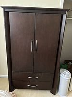 Front view of dark brown wood armoire with closed doors and two drawers underneath, silver handles.