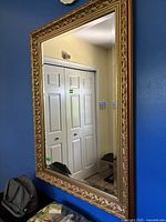 Large gold Baroque style framed wall mirror mounted on blue wall showing reflective surface and detailed ornate edges.