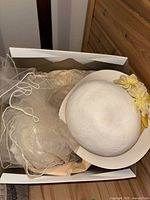 White wide-brim hat with yellow ribbon and artificial yellow flowers, shown in storage box with tissue paper and attached sheer veil visible.