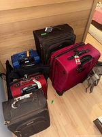 Six pieces of varying sized soft luggage in black, maroon/red, blue, brown, and red colors, some with carry handles and wheels, arranged on the floor