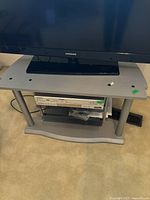 Metal and glass TV stand holding a Samsung television, grey top and bottom platforms with metal legs, glass shelves in between.