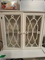 Front view of mini off white cabinet with two glass doors having decorative wooden lattice design and metal knobs.